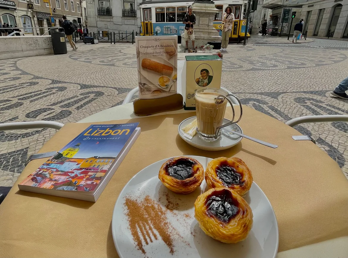 Three pastéis de nata dusted with cinnamon and a milky coffee on a café table, with a tram on the patterned pavement behind