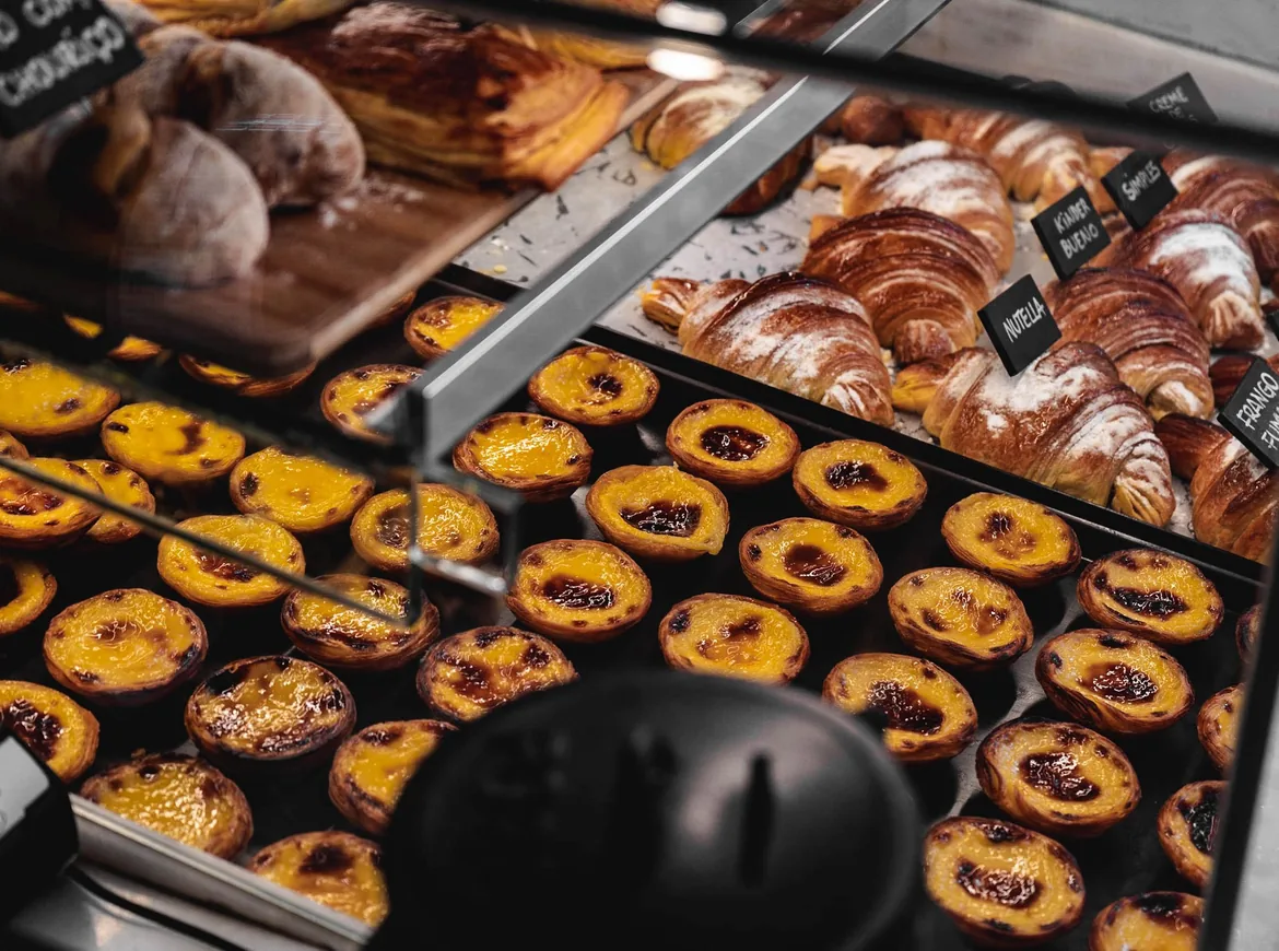 Trays of custard tarts and croissants behind the glass of a Lisbon bakery counter, handwritten labels above them
