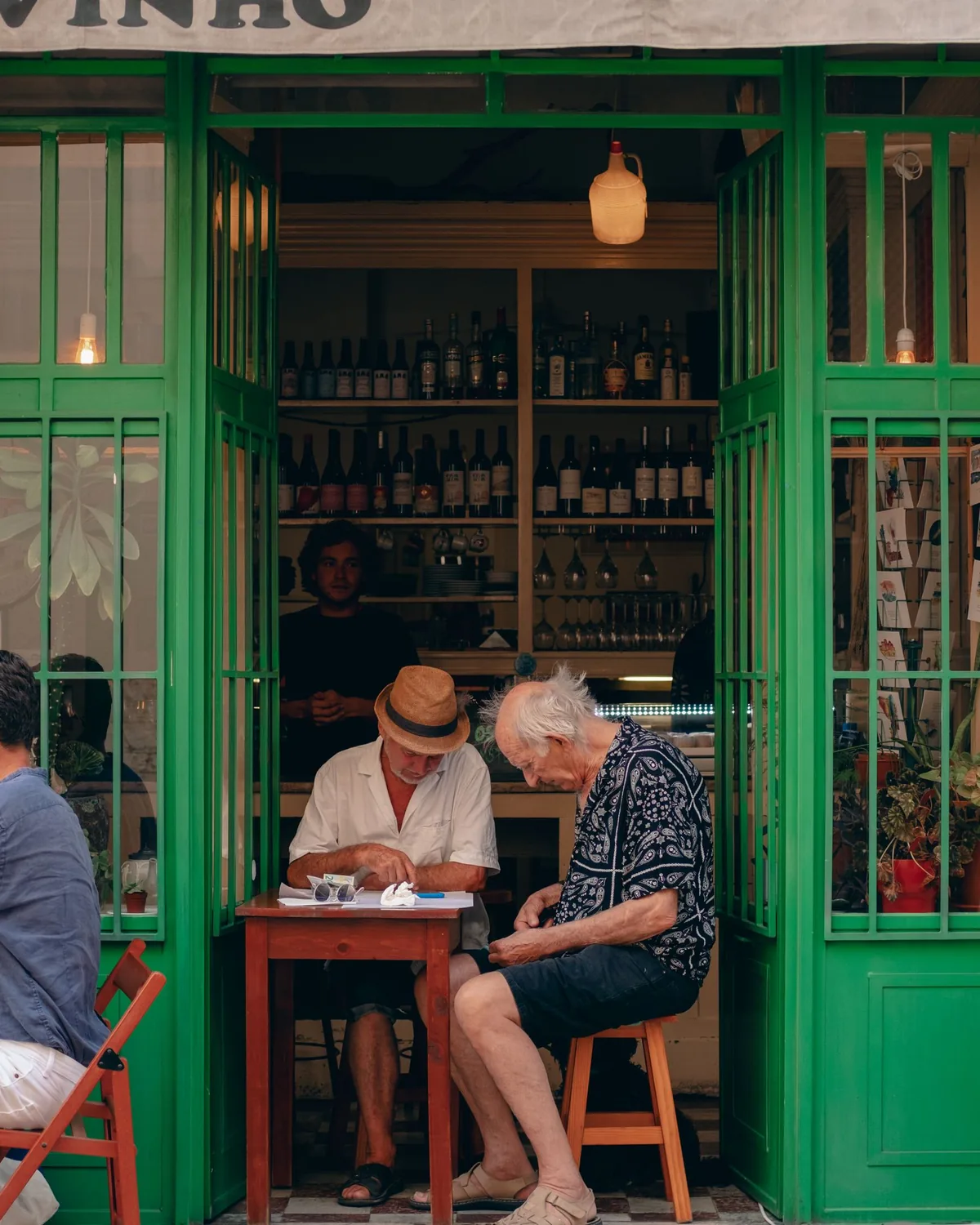 Three men at a small table in the open green-framed doorway of a Lisbon wine bar, bottles on the shelves behind them